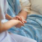 A nurse sitting on a hospital bed next to an older woman holding her hand.