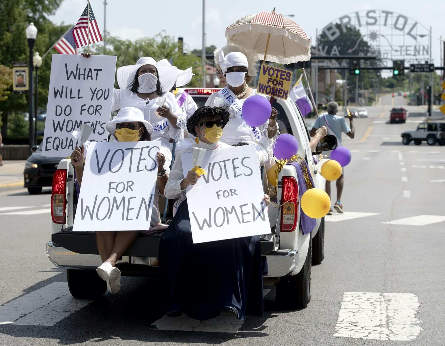 Women in all white sitting in the back of a truck with "votes for women" signs.