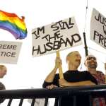 Three people stand with signs supporting marriage equality as the LGBTQ flag waves in the background.