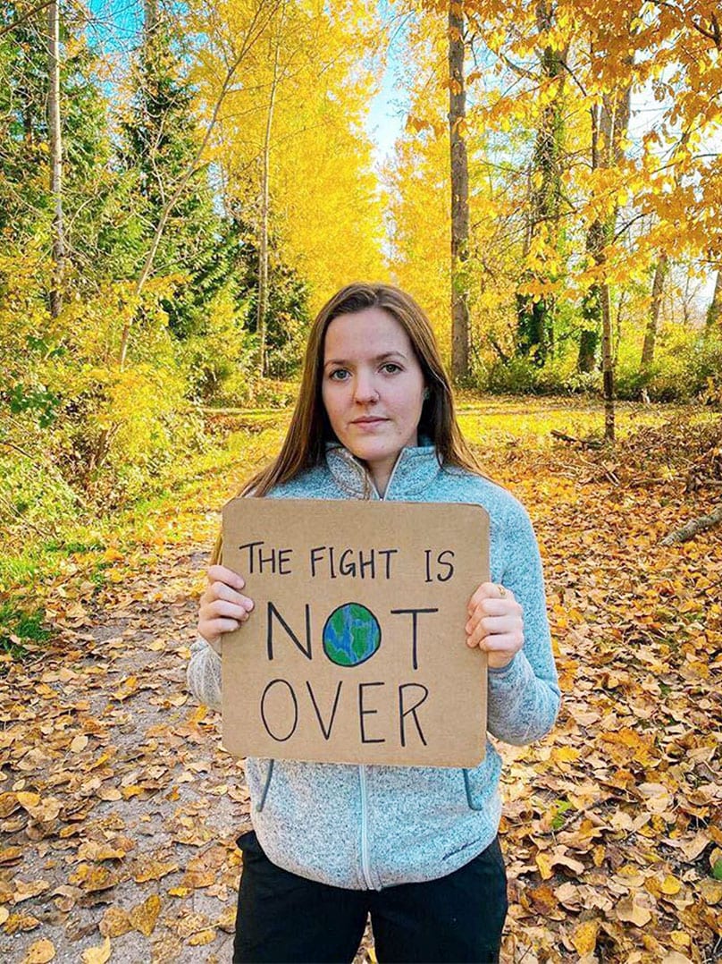 A woman stands in a forest with a sign that reads, "The fight is not over."