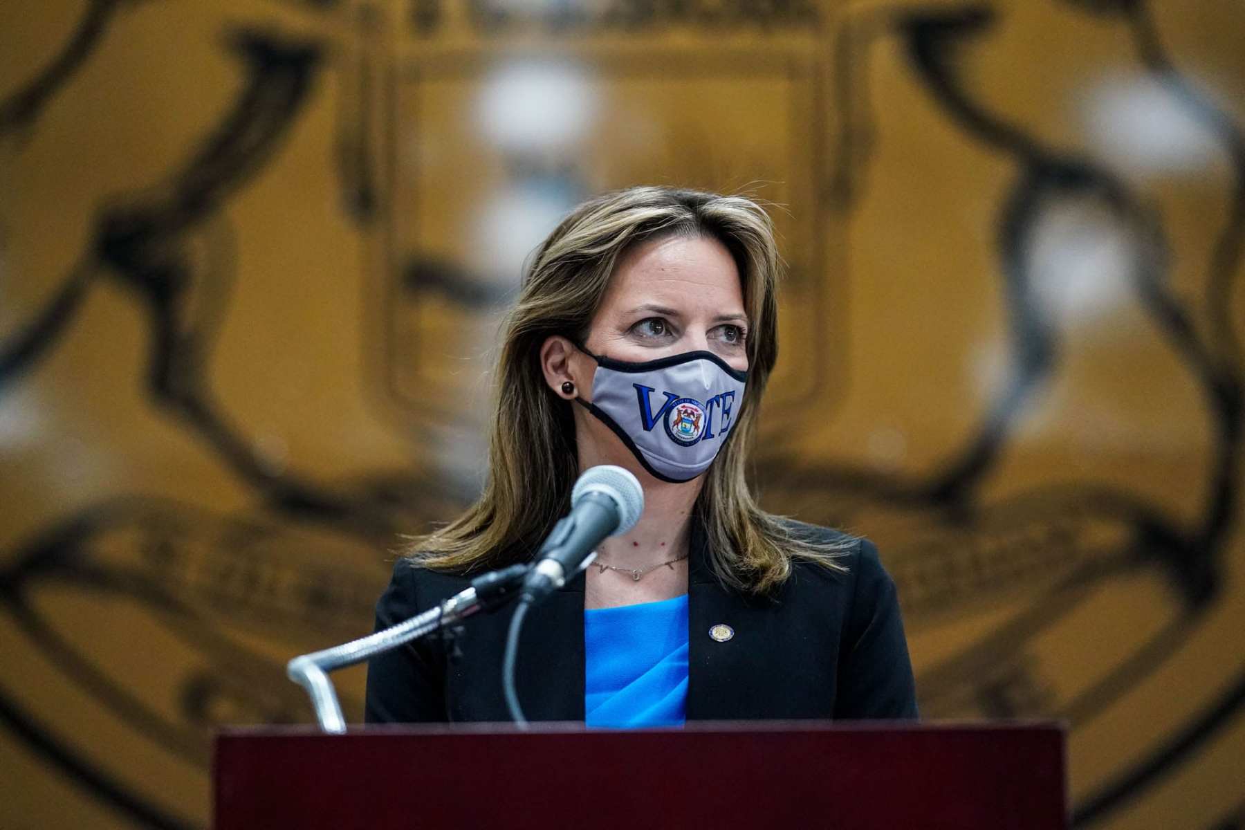 Michigan Secretary of State Jocelyn Benson stands at a podium with a mask on that says "vote."