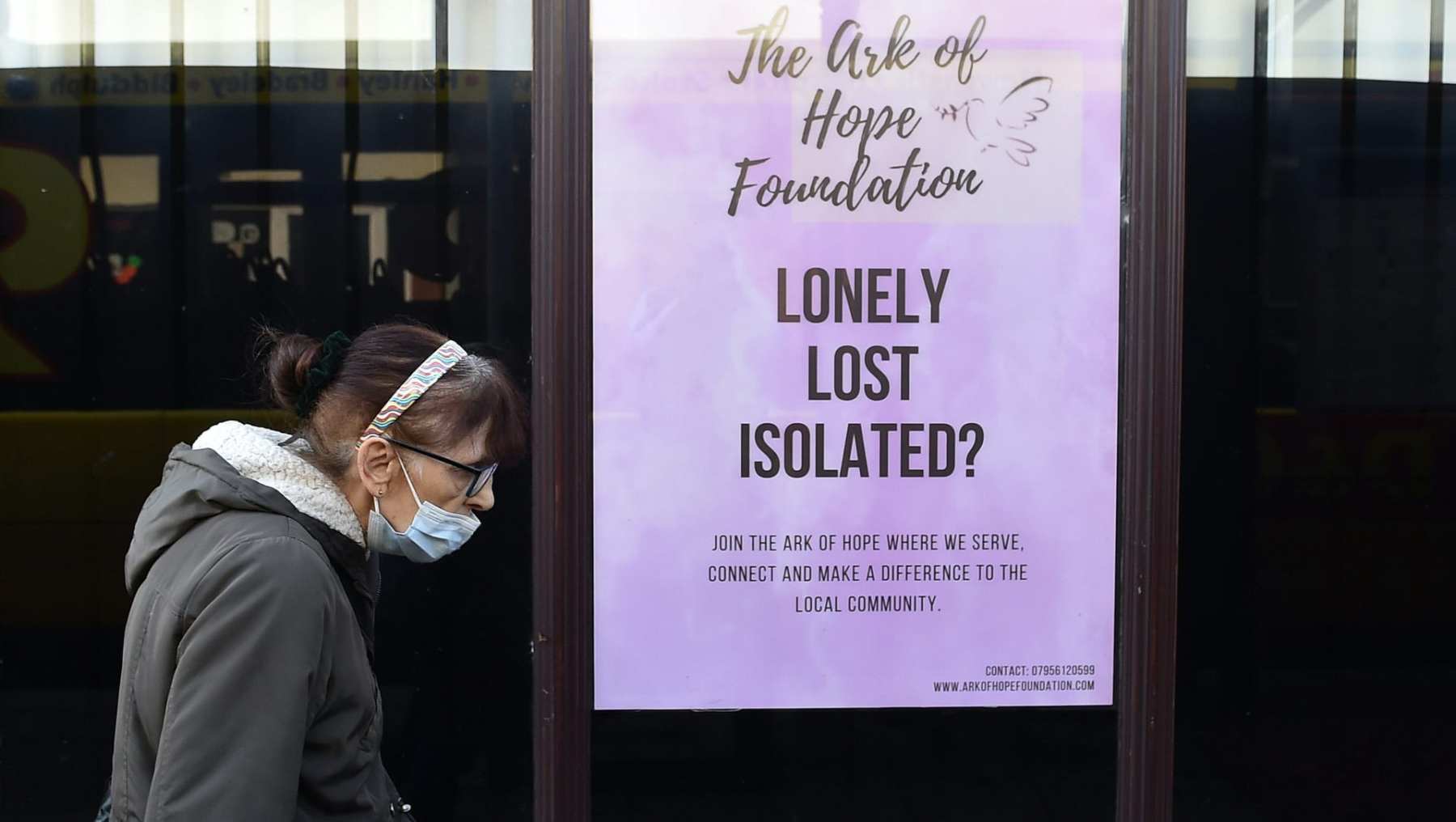 A woman wearing a mask walks past a sign that reads "Lonely, lost, isolated?"
