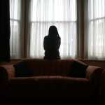 A young Asian woman suffering from domestic violence stands alone in the bay window of her home.