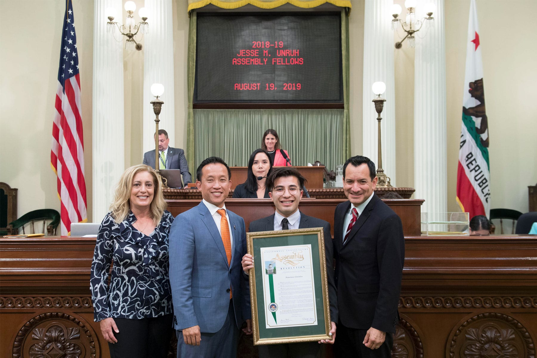 Severiano Christian standing in front of the chamber podium holding am assembly certificate with fellow assembly members.