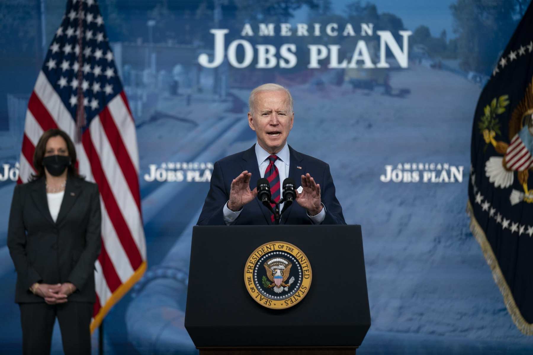 Joe Biden speaking at a podium in front of a backdrop that reads "American Jobs Plan"