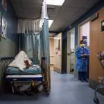 A patient rests in a corridor as a nurse looks on in the distance.