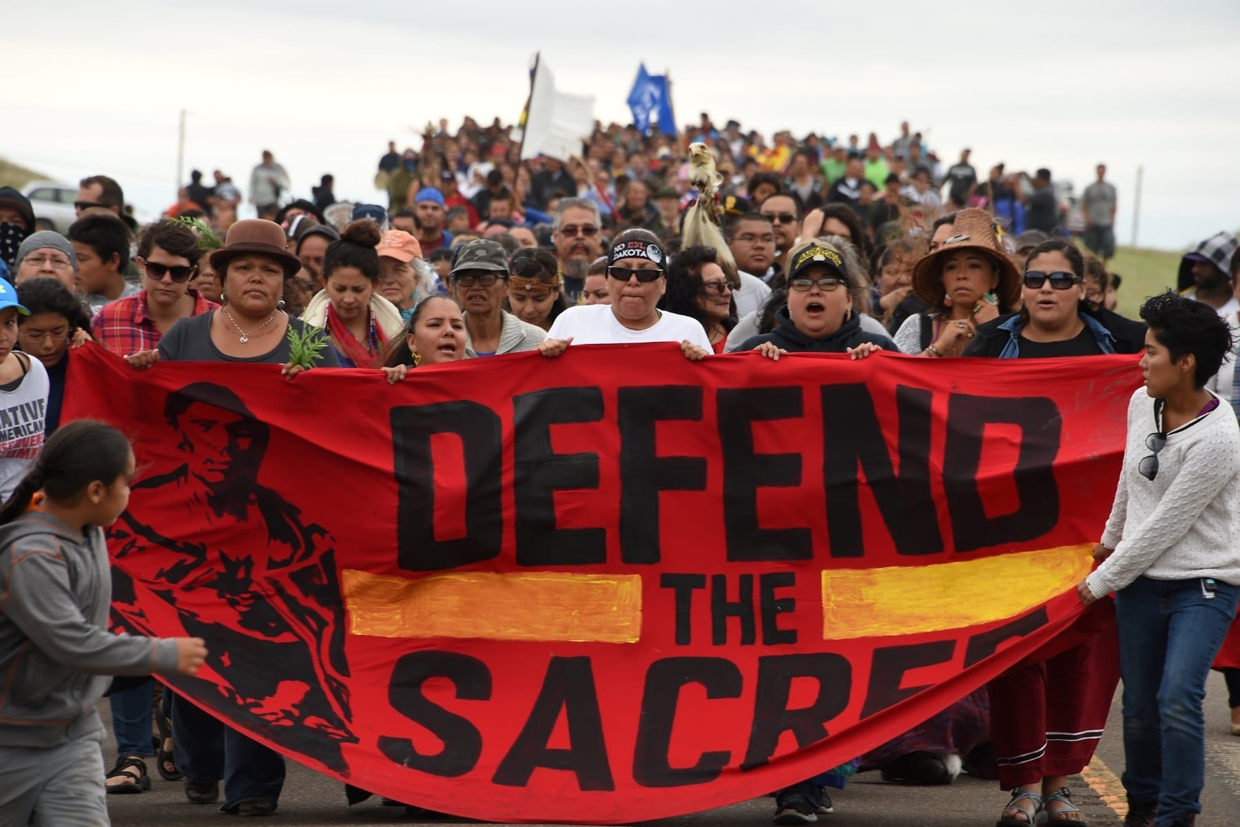 A group of Native American protesters marching with a sign that says "Defend the Sacred."
