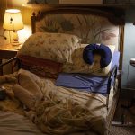 The bedroom of an elderly person is pictured. The bedsheets are undone and on the bed rests a neck pillow and a box of tissue paper that appears empty.