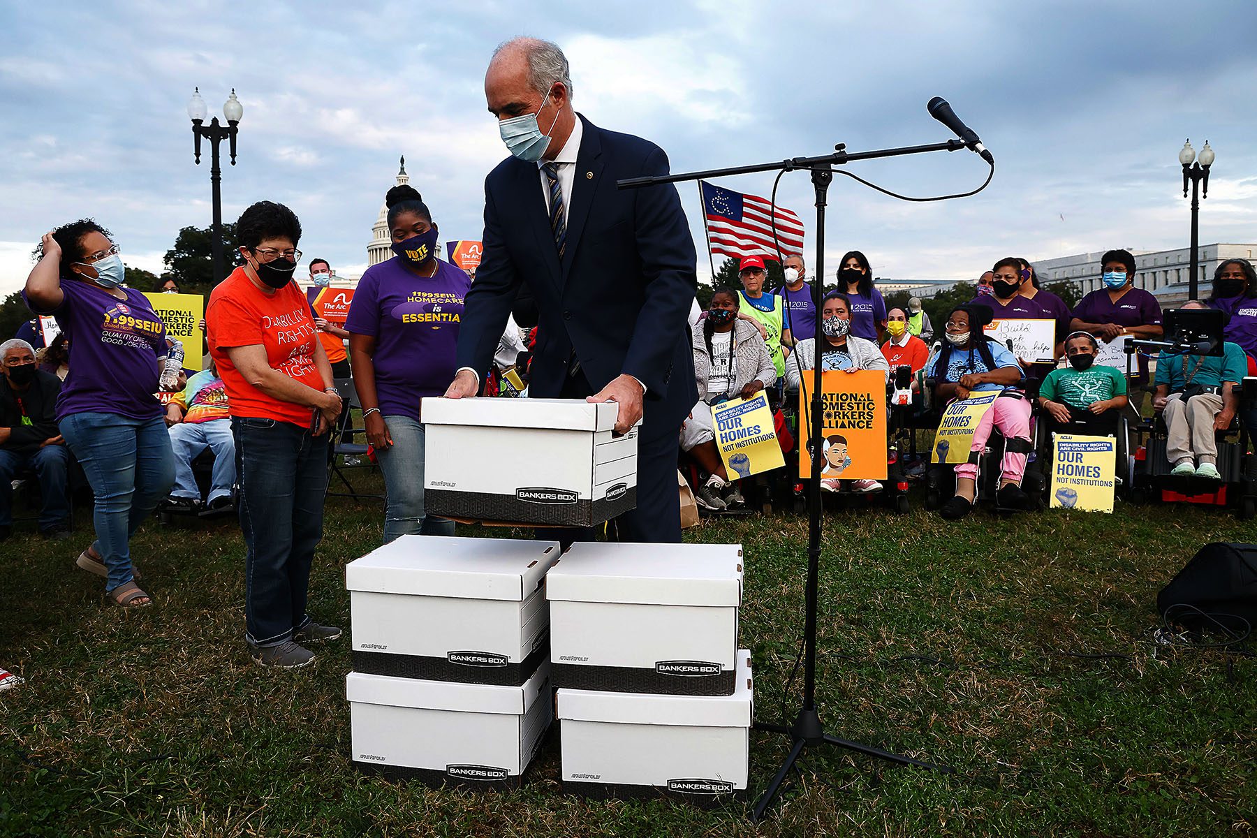 Bob Casey lifts a large white cardboard box while activists watch all around him. The Capitol building is seen in the background.