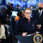 Deanne Criswell speaks at a podium on which is the presidential seal is hung. Joe Biden can be seen listening behind her.