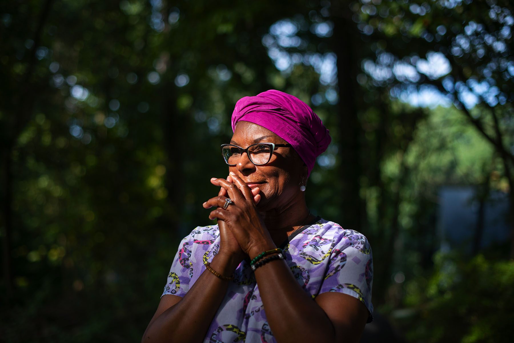 A woman wearing a pink turban and purple scrubs's face is lit by a ray of sun. She is surrounded by trees.