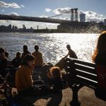 A woman accompanied by two children is on the phone as people sit near the East River. The manhattan skyline is seen in the background.