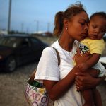 A young woman presses her face against a young child's face while standing on rubble road where cars are parked.