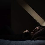 A woman lays on a bed as soft light trickles into the room from a skylight.