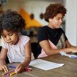 A child and woman sit together at a table while the child colors and the mother works on a laptop.