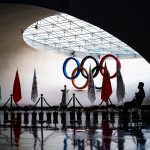 A man guards the olympic flame, which stands in front of Olympic rings.