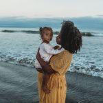 A pregnant mom and her daughter embrace near the ocean.