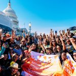 A large group of people hold their fists up in the air and smile in front of the Congress building. They are wearing t-shirts that read 