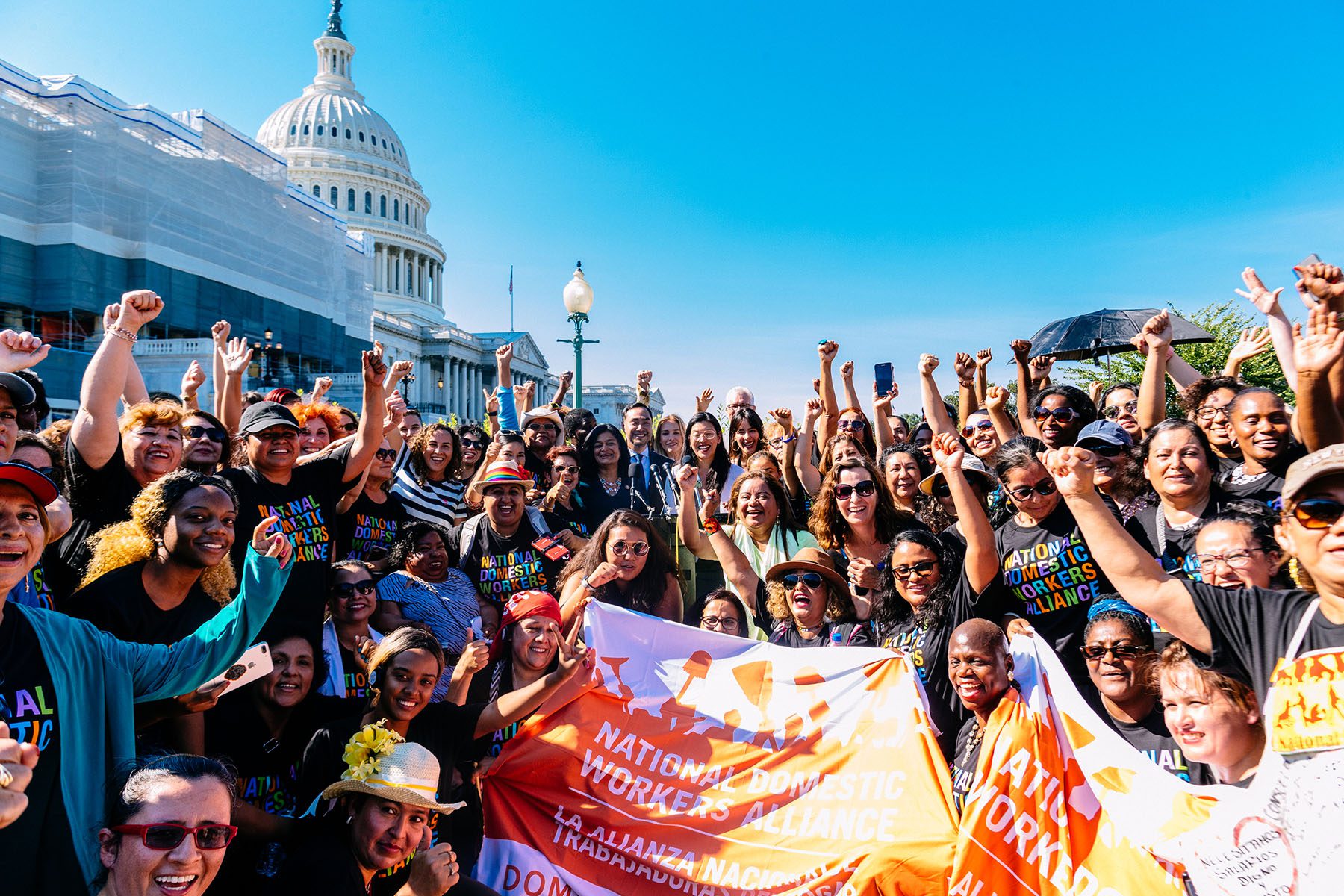 A large group of people hold their fists up in the air and smile in front of the Congress building. They are wearing t-shirts that read "National Domestic Workers Alliance."