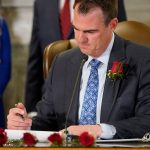 Kevin Stitt sits at a desk and signs into law a bill criminalizing abortions. He is wearing a red rose on his lapel. Several other red roses are seen on his desk.