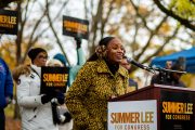 Summer Lee is seen smiling at a podium as people hold signs that read 