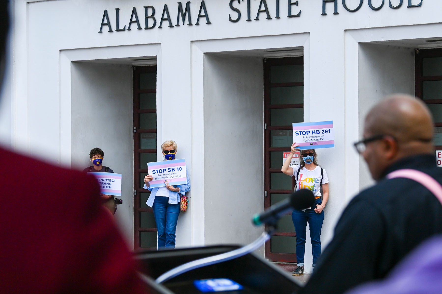 Several people hold up signs that read "STOP HB 391 Anti-Transgender Youth Athlete Bill," and "STOP SB 10 Anti-transgender Youth Medical Care Bill" during a rally in front of the Alabama State House. A person is seen making a speech at a podium in the foreground.