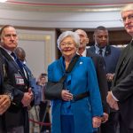 Gov. Kay Ivey wears a sling around her arm as she smiles before walking into the chamber. She is surrounded by several men.