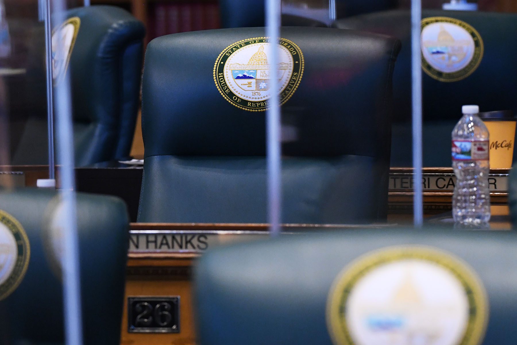 Empty desks and desk chairs are seen at the Colorado State Capitol. The chair are engraved with a "State of Colorado, House of Representatives" emblem.