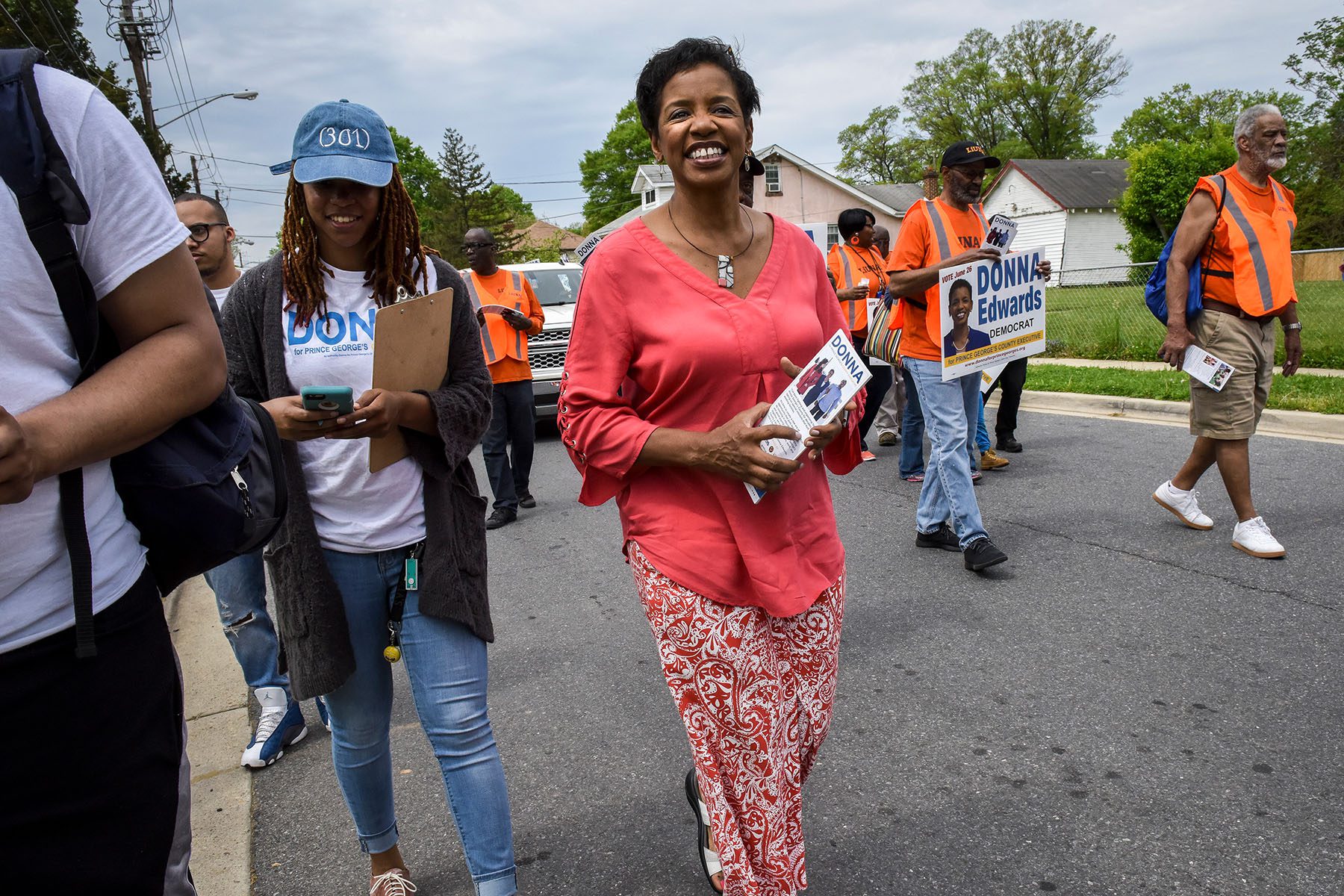Donna Edwards walks down a street carrying a small pamphlet. Others around her carry signs that read "Donna Edwards Democrat" and wear shirts that say "Donna for Prince George's."