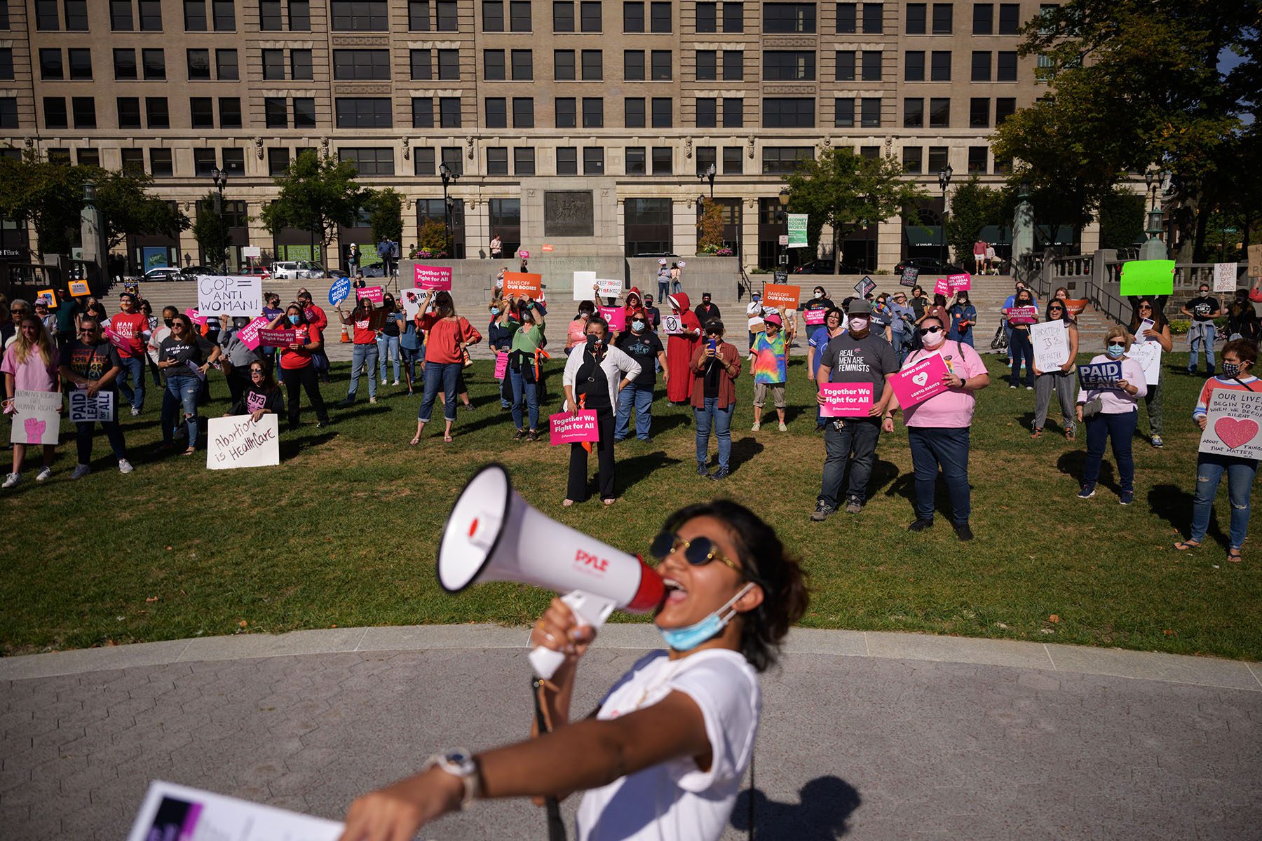 A protesters speaks into a megaphone in the foreground. In the background, people hold signs that read "Together we fight for all" and "Abortion is healthcare."