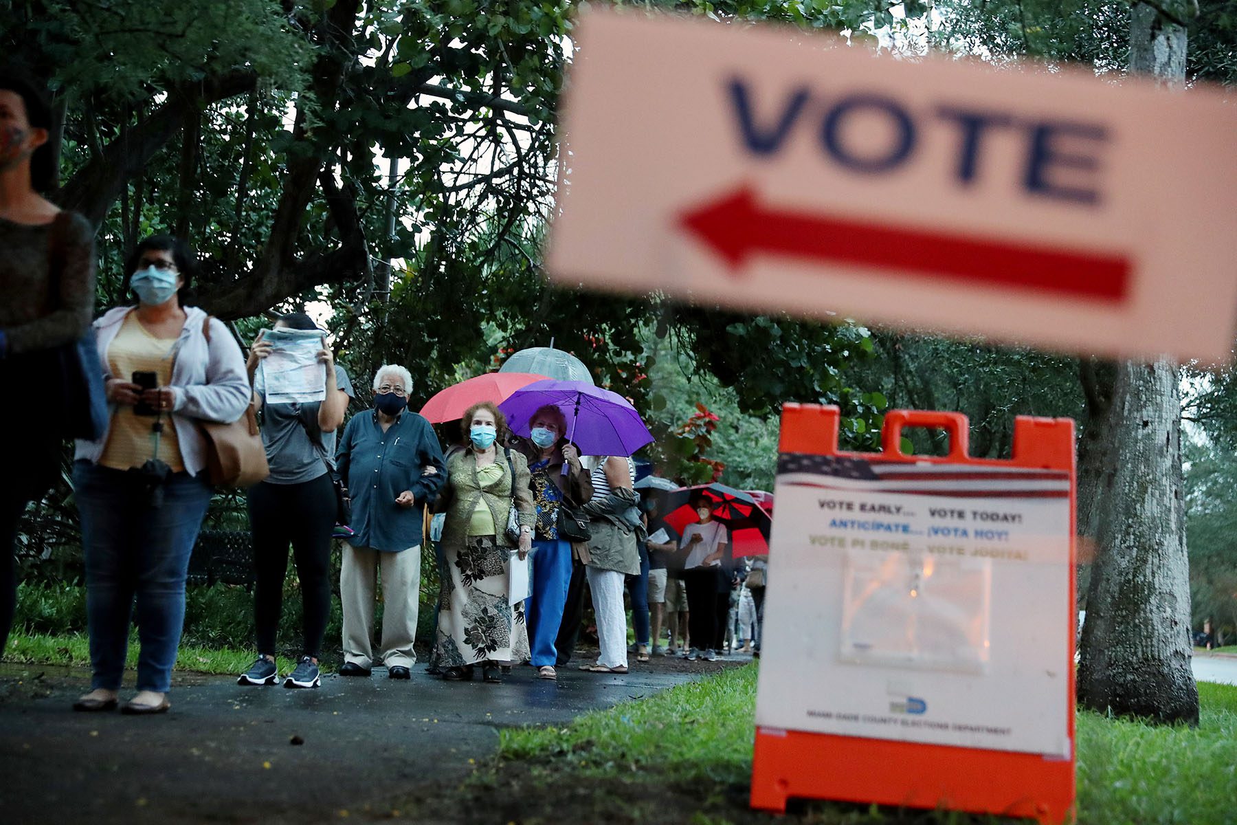 Voters wait in line to cast their ballots on a rainy florida morning. Several people in line are holding umbrellas and wearing face masks. A "vote" sign is seen blurred in the foreground.