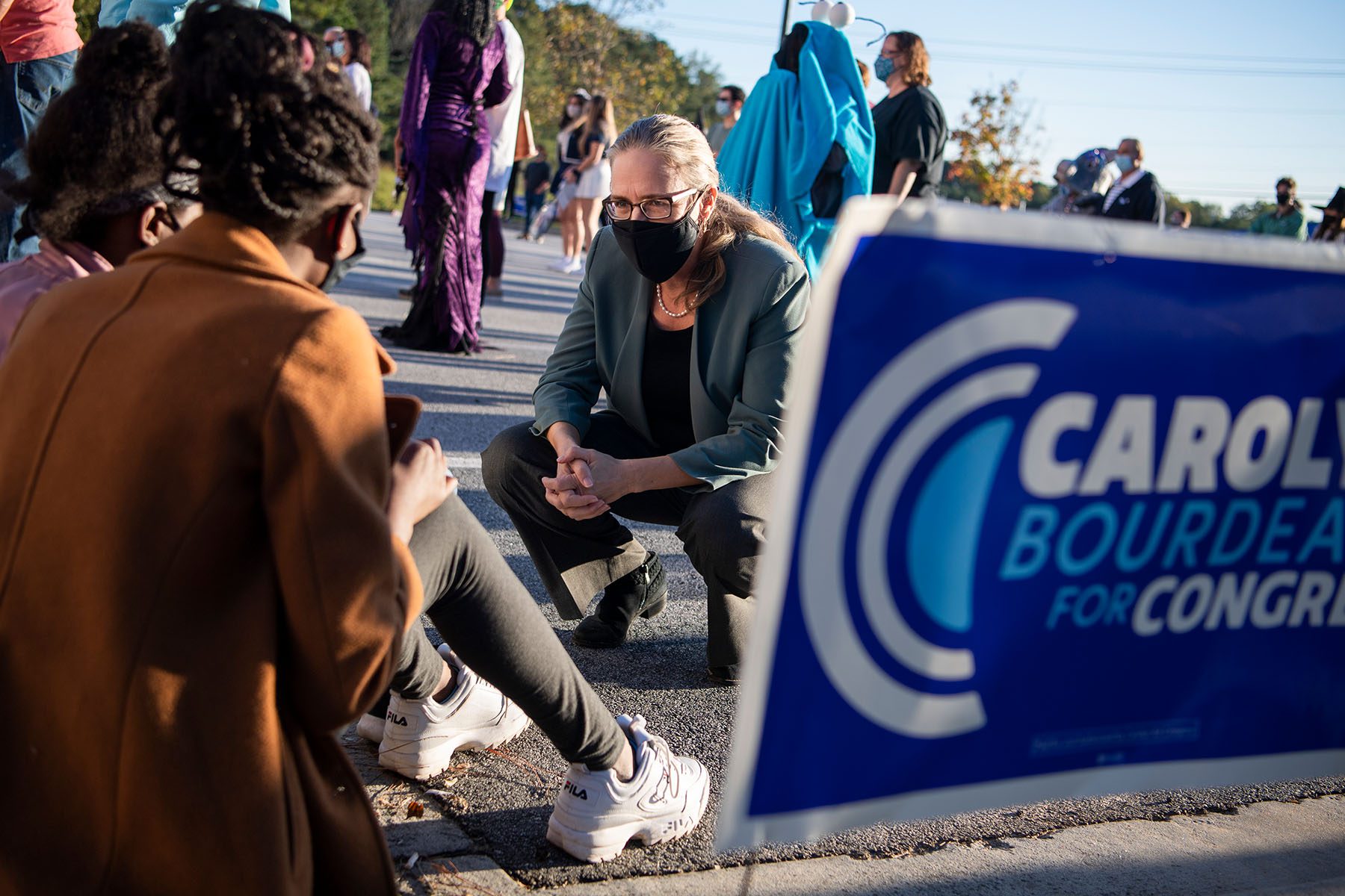 Carolyn Bourdeaux kneels to speak to supporters sitting on a curb. A sign that reads "Carolyn Bourdeaux for Congress" is seen in the foreground.
