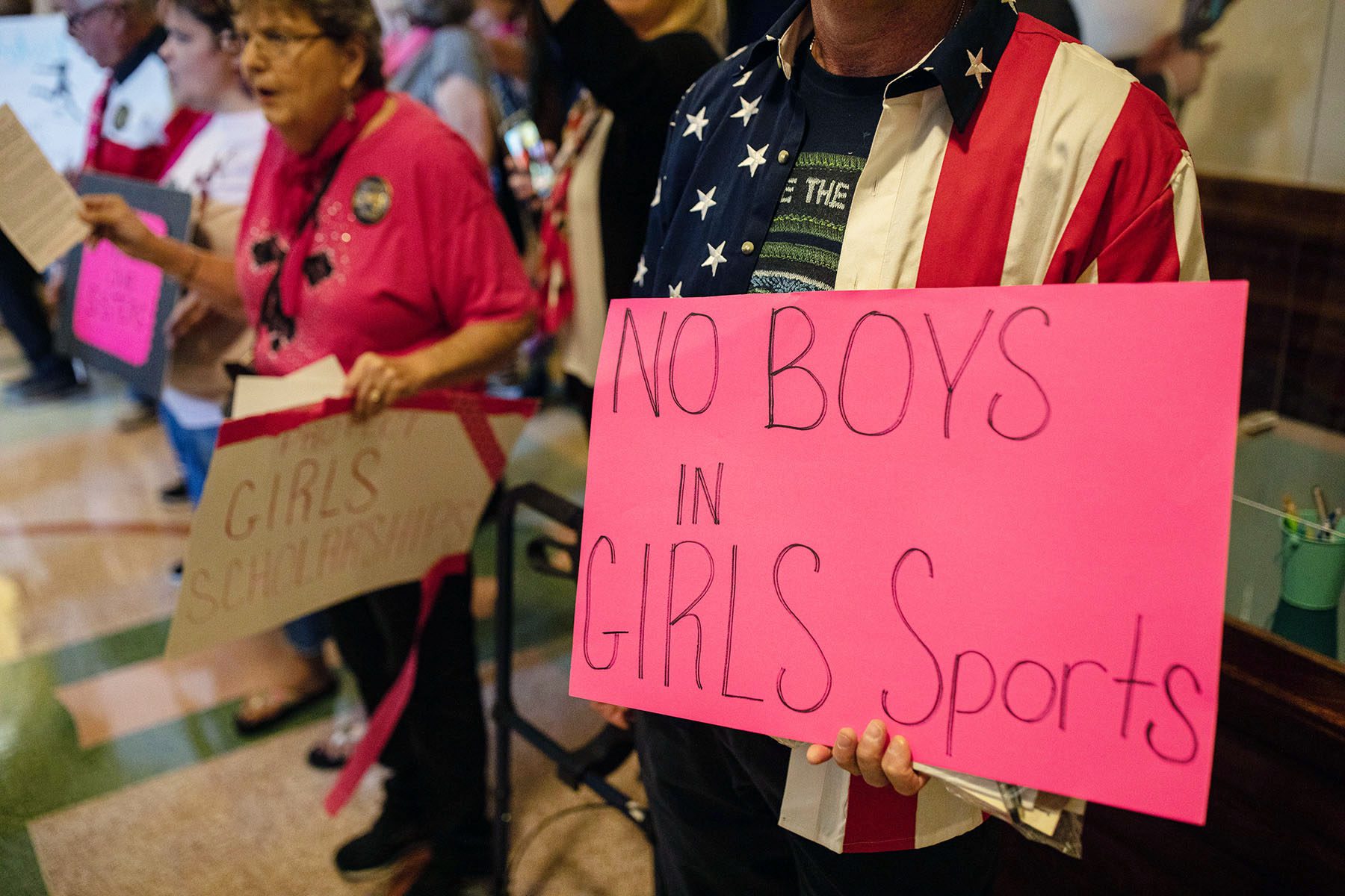 A man wearing an american flag button down holds a sign that reads "no boys in girls sports."
