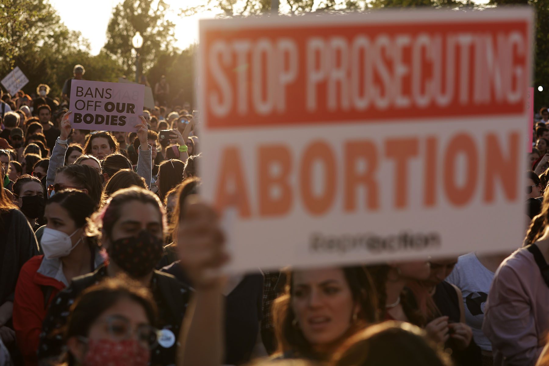 A large crowd of people protest in front of the Supreme Court. A sign in the foreground reads "Stop Prosecuting Abortion."