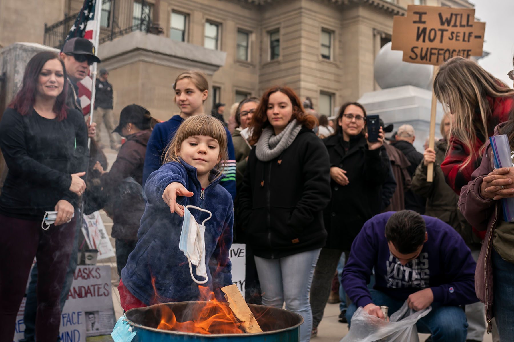 A child tosses a surgical mask into a fire. The child is surrounded by demonstrators holding American flags and signs, one of which reads "I will not self-suffocate."