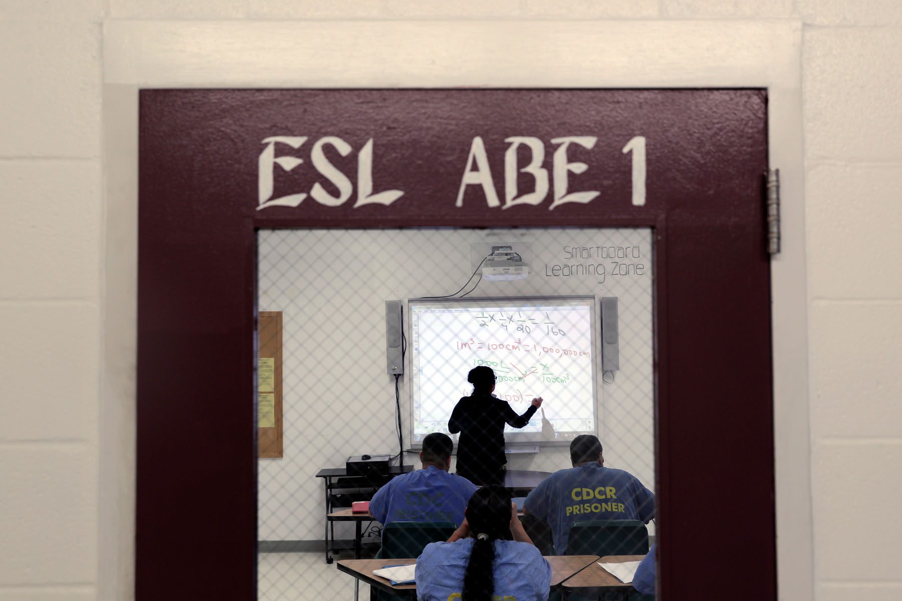 Prisoners are seen studying inside a class room. The teacher is resolving a math problem on the whiteboard. The door of the classroom reads "ESL ABE 1."
