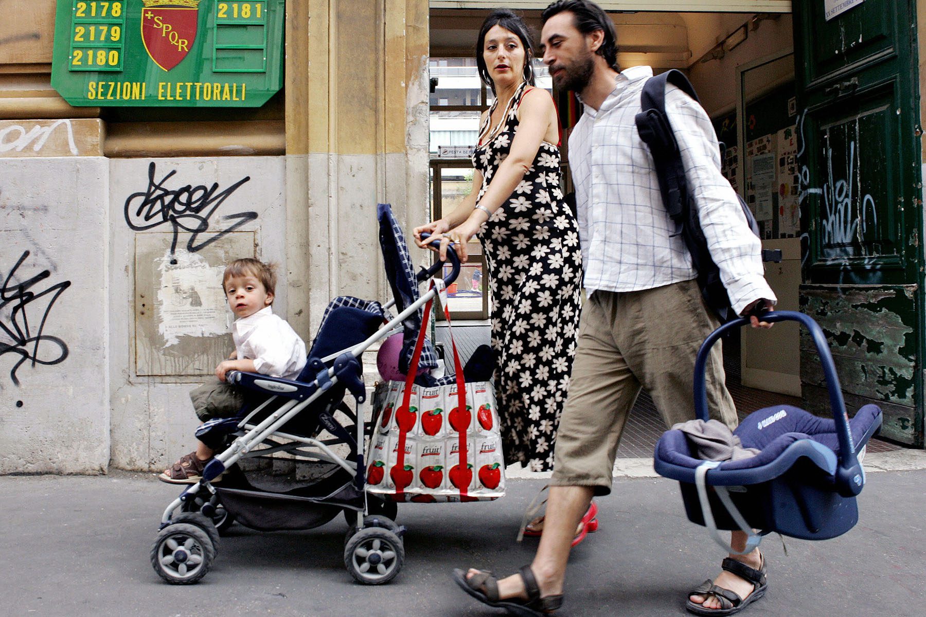 A woman pushes a stroller with a small child on board as she speaks to a man holding a baby carrier.
