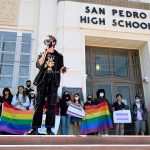 Dylan Keener holds a microphone while speaking in front of San Pedro High School. Behind him, students hold pride flag.