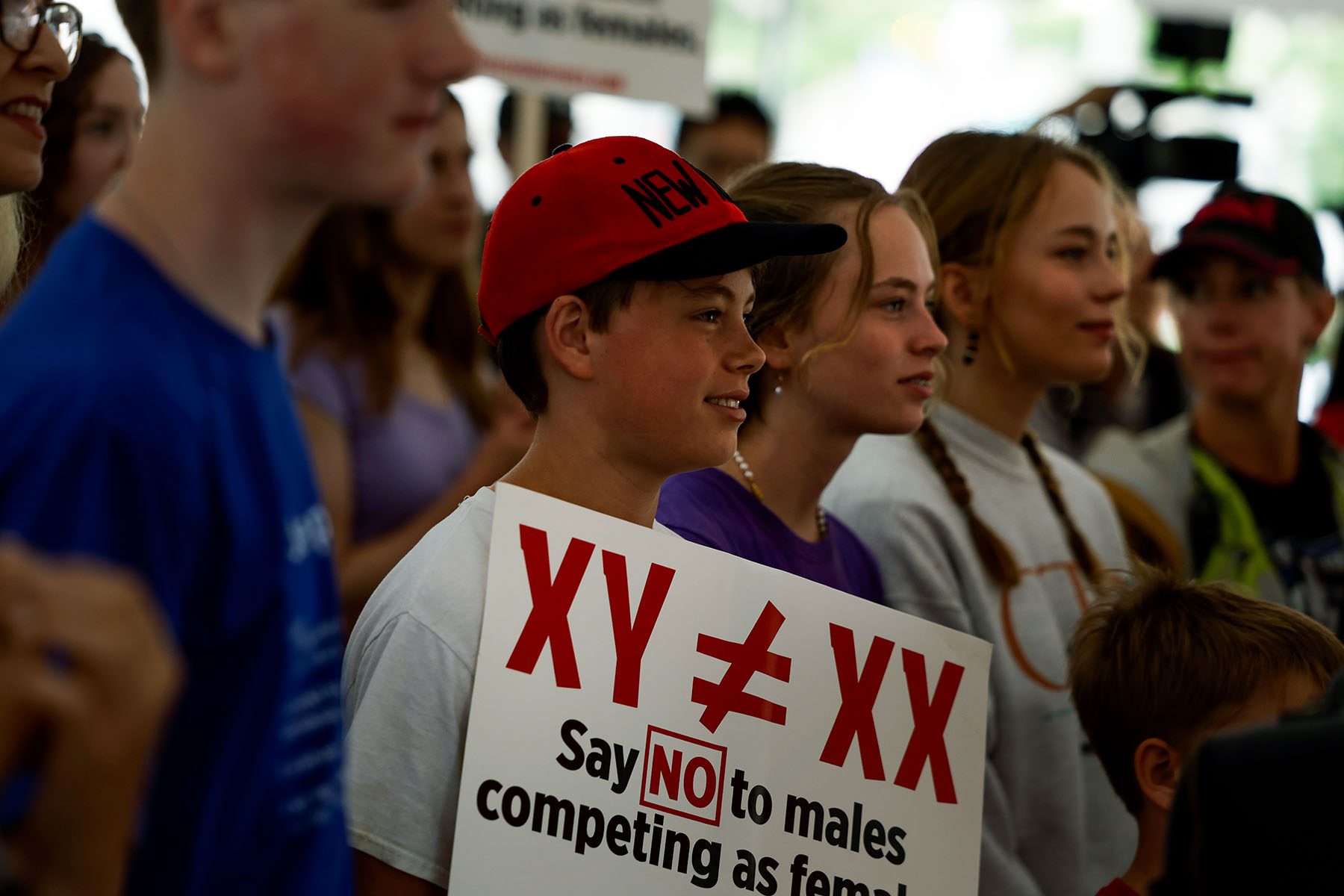 Demonstrators listen during a rally. In the foreground, a young person holds a sign that reads "XY is not equal to XX, say no to males competing as females"