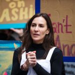 Alessandra Biaggi speaks as people holding signs demonstrate behind her.