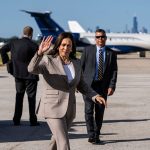 Vice President Kamala Harris waves to the press before boarding Air Force Two. Behind her are two secret service agent and a plane.