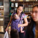 A mother with a baby in a carrier on her chest shops for groceries.