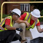 A woman and man work together to install solar panels in construction gear.