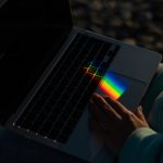 A person uses a laptop on which light is refracted, making a small rainbow appear on the touchpad.