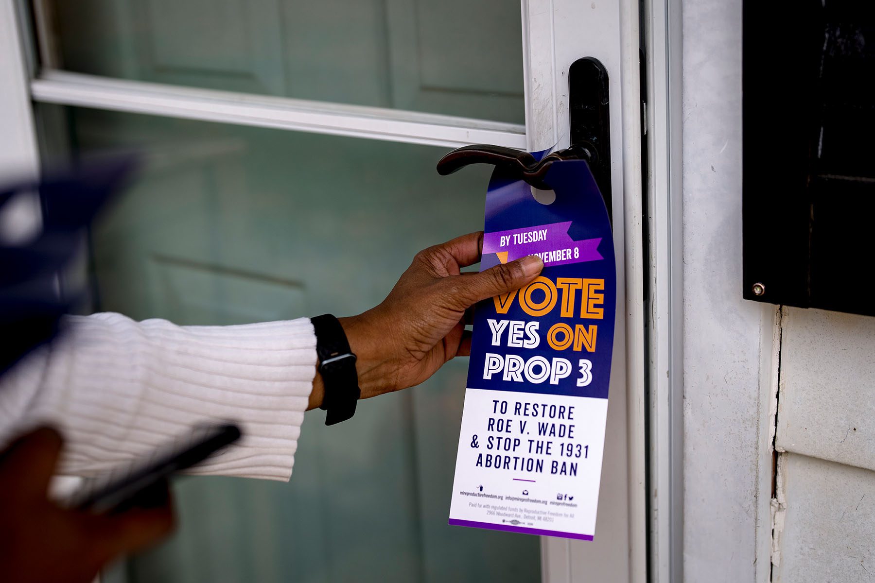 A volunteer places a door tag in support of Proposal 3 on a door. The tag read "vote yes on prop 3 to restore roe v. wade and stop the 1931 abortion ban."