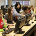 A woman stands and teaches students in a room full of computers.