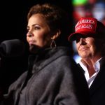 Kari Lake speaks into a microphone as President Trump looks at her smiling at a rally.