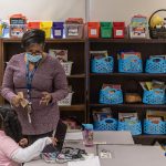 A second grade teacher wearing a face mask instructs one of her students in a colorful classroom.