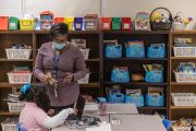 A second grade teacher wearing a face mask instructs one of her students in a colorful classroom.