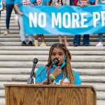 Tamia Cenance speaks at a podium as activists hold a banner that reads 